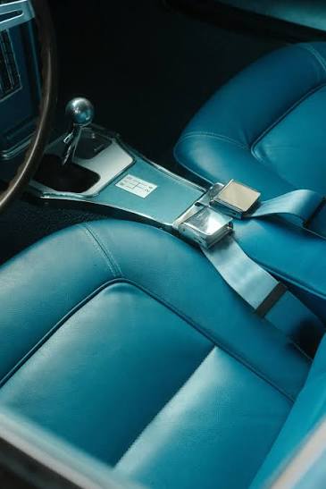 Interior of a classic car with teal blue leather seats and a chrome lap belt buckle fastened across the centre of the bench.