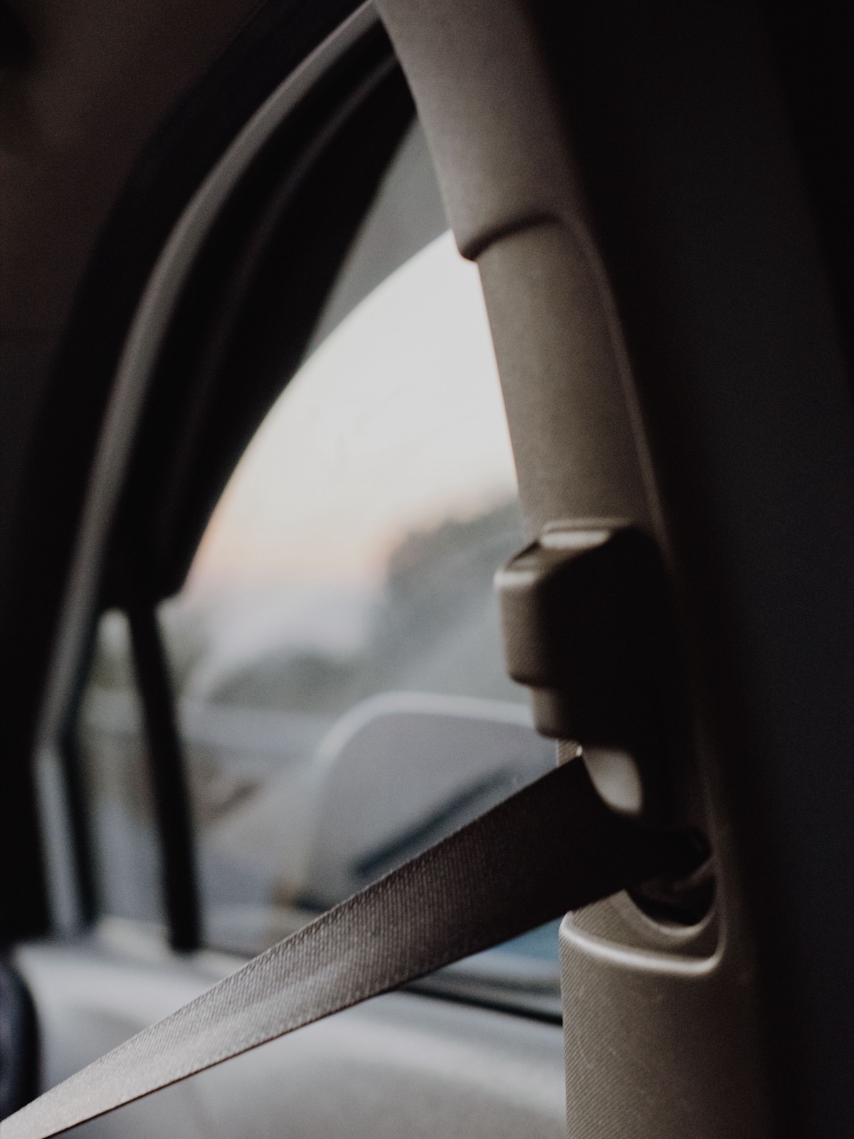 Seat belt webbing running through an upper anchorage point on a vehicle B-pillar, photographed against a window at dusk.