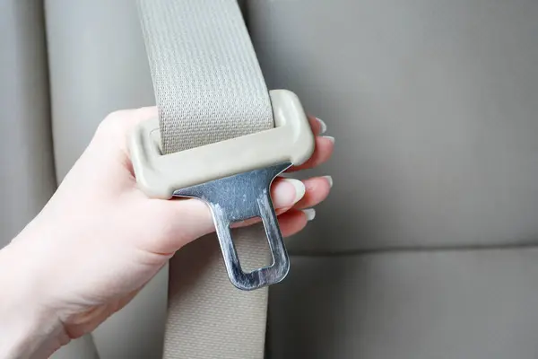 A hand holding a beige seat belt tongue plate away from the seat so the metal latch profile is clearly visible.